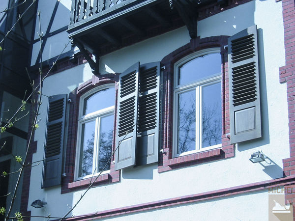 Zwei hohe Fenster mit braunen Holzfensterläden an einer Fassade im Altbau-Stil.