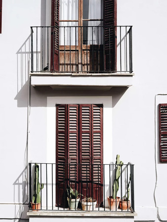Balkon mit schwarzen Geländern, Holztüren und Blumentöpfen mit Kakteen an weißer Fassade.