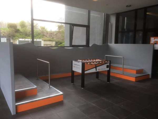 Moderner Aufenthaltsbereich mit Tischkicker, großen Fenstern und orange-grauen Sitzstufen.